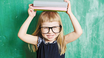 A little girl with glasses holding a stack of books on her head.